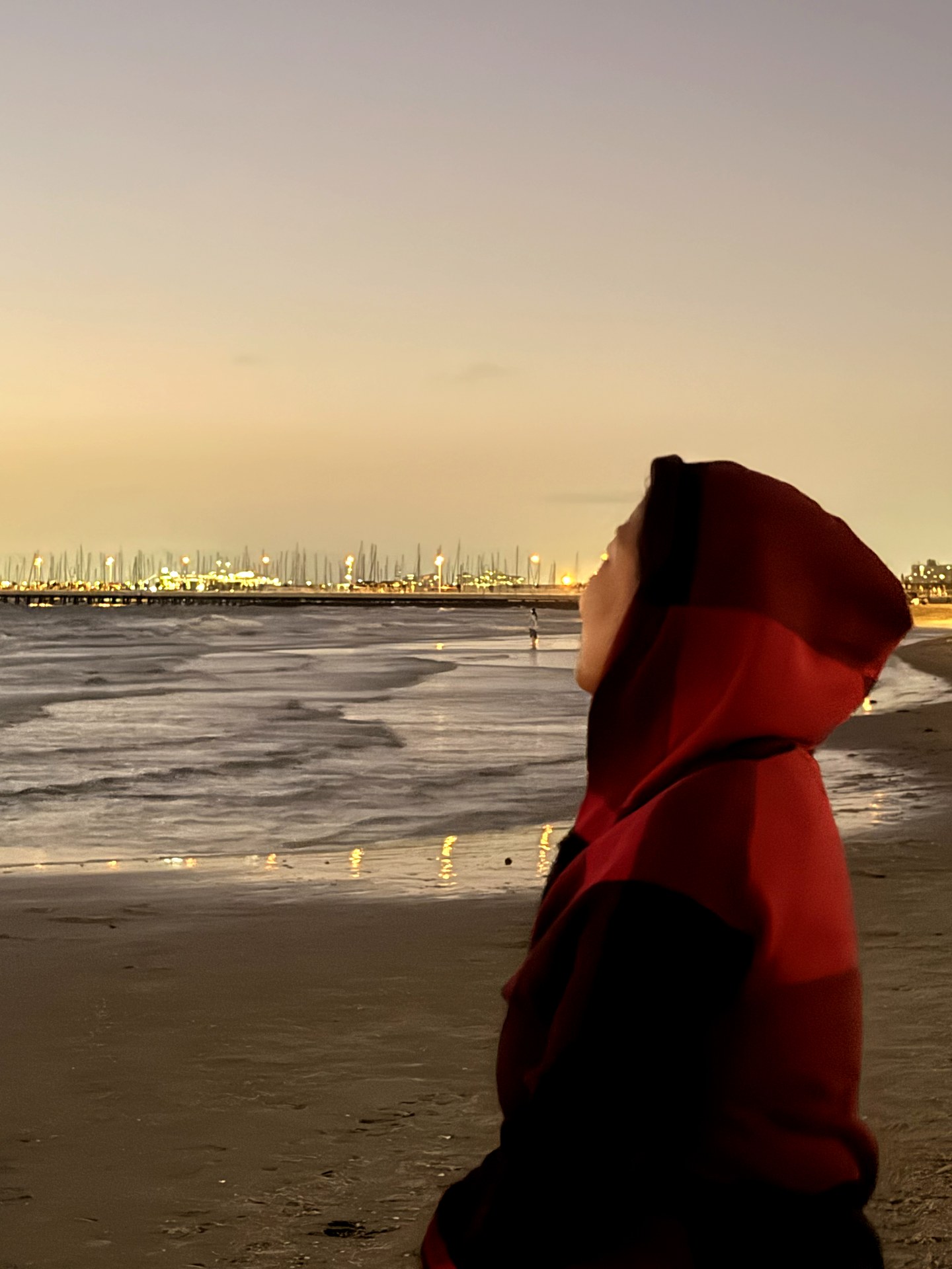 A person in a red-and-black hood beside the water at St Kilda, with marina lights beyond